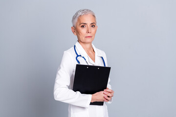 Poster - Photo of attractive senior woman hospital doctor worker hold paperholder dressed white robe uniform isolated on gray color background