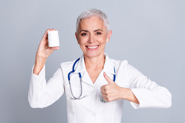 Sticker - Portrait of aged woman professional doctor thumb up pills wear white coat isolated on grey color background