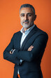 © wolfhound911 - A confident middle-aged Arabian man in a tailored suit, arms crossed against a vibrant orange background, projecting authority and determination.