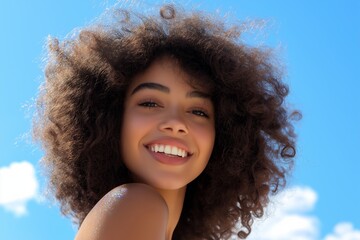 A woman with curly hair is smiling and looking at the camera. The image has a bright and cheerful mood, with the blue sky and the woman's happy expression