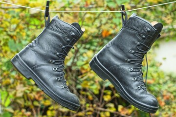 pair of military protection  leather high stylish black lace up army rubber sole modern new heavy duty mens fashion boots hanging on clothespins in green garden in daytime