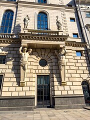 A close-up shot of a grand building facade in bright sunlight, featuring two muscular male sculptures supporting a balcony. The image captures intricate architectural details, emphasizing the strength