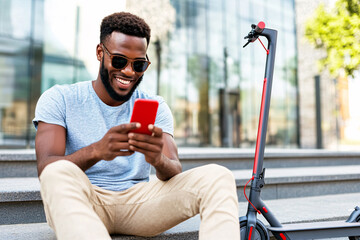 A smiling African American man is sitting outdoors on concrete steps, holding a red smartphone. Dressed casually, he is relaxed next to an electric scooter in an urban environment..