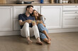 © fizkes - Positive dreamy married couple resting on warm floor in modern kitchen, sitting close, looking away with happy smiles, talking, discussing future plans, drinking hot beverage