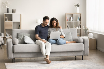 Wall Mural - Millennial couple using Internet service, application on laptop, sitting on couch together, watching online content, enjoying leisure, wireless technology in comfortable home interior