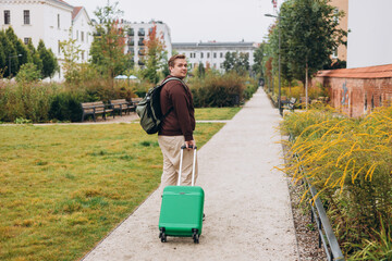 Happy 30s man pulling green suitcase outdoors. Rear view. Copy space. Man walking on the street with travel bag.