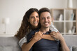 © fizkes - Happy beautiful young loving wife hugging cheerful handsome husband from behind. Positive married couple enjoying being at home, looking at camera with toothy smiles, posing on couch for portrait