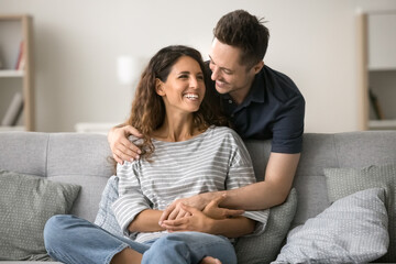 Wall Mural - Cheerful attractive couple in love enjoying leisure at home, hugging at comfortable couch, talking, laughing. Happy handsome husband embracing wife from behind