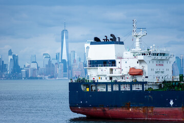 Wall Mural - The ship's stern takes focus as it glides through the bay, with New York City's skyline blurred in the background. The cloudy sky casts a soft, steel-blue tone over the entire scene.