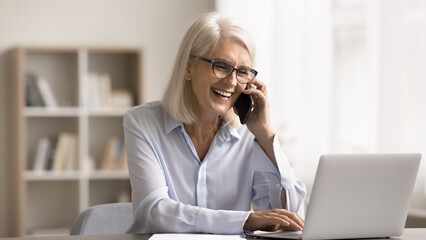 Canvas Print - Happy businesswoman typing on laptop keyboard and talking on smartphone, working seated at workplace desk in office, enjoy pleasant business conversation, share great news by phonecall. Communication