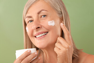 Canvas Print - Senior woman applying face cream on green background, closeup