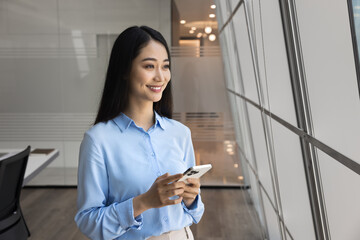 Cheerful beautiful young Asian manager woman holding mobile phone, standing in modern office space, looking at window away, thinking on Internet communication, business success