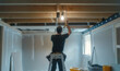 © Michael - A male electrician in uniform installing light fitting in new apartment, standing on a ladder