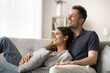 © fizkes - Dreamy cheerful young married couple resting on couch, looking away, thinking, dreaming, planning vacation, laughing, talking, discussing new home apartment, mortgage, enjoying leisure