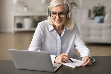 Aged woman wear headphones engaged in online study using laptop, participating in video call or virtual meeting, taking notes. Teacher lead on-line lesson, explain topic to learner. Tuition, education