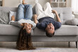 © fizkes - Cheerful cute young couple relaxing on grey soft couch with legs on back and hanging heads to floor, lying upside down, talking, laughing, smiling, having fun at home, enjoying funny leisure