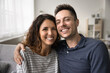 © fizkes - Cheerful attractive couple head shot video call portrait. Happy handsome man and pretty woman sitting on home couch, hugging with head touches, looking at camera with toothy smiles