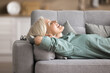 © fizkes - Side view serene older woman take daytime nap lying on cozy sofa in modern living room, smile, while breathing fresh conditioned air inside, spend free time alone at home. Rest, break, fatigue relief