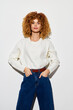 © SHOTPRIME STUDIO - Fashionable young woman with curly hair, wearing a cream sweater and blue jeans, standing confidently against a plain white background