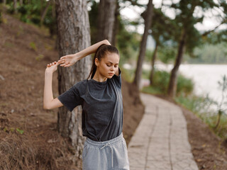 Relaxed young woman stretching outdoors by the serene lake surrounded by trees, enjoying peaceful nature and personal wellness in casual gray attire