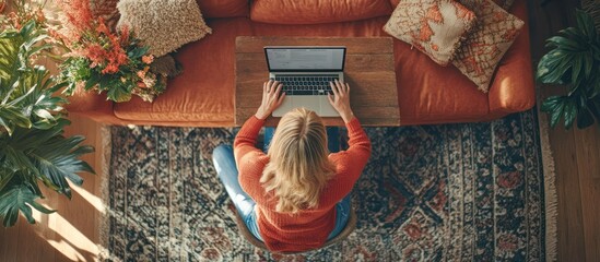 Canvas Print - Woman working on laptop from home, sitting on a rug, with orange couch, plants, and sunlight coming through the window.