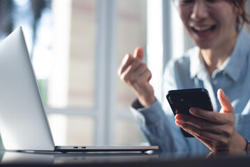 Wall Mural - Cheerful young asian business woman using mobile phone during working on laptop computer, online meeting in office. Casual business woman, entrepreneur smiling during video calling via smartphone