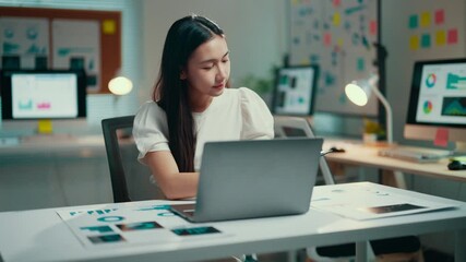 Aufkleber - Young asian businesswoman sits at her modern office desk, working late into the night on her laptop. Focused and determined, she navigates through data and strategies
