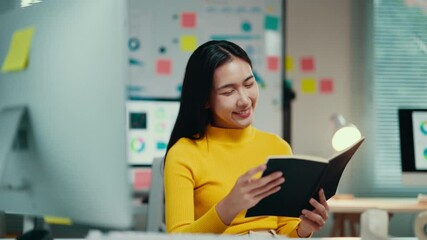 Poster - Asian businesswoman smiles while reading a book at her office desk. Dressed in a yellow sweater, she enjoys a moment of relaxation amidst her professional environment