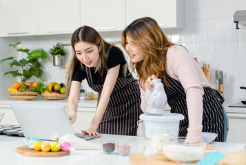 Two happy young Asian female friends, woman wearing aprons, cooking dough baking cakes, dessert, looking online recipe guidebook on laptop computer enjoy weekends in kitchen, teenage lifestyle at home