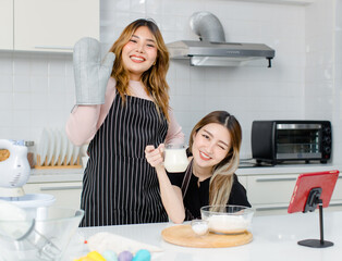 Two happy young beautiful Asian female, woman looking at camera smiling wearing aprons enjoying fun while making bakery in kitchen concept of teenage lifestyle at home.