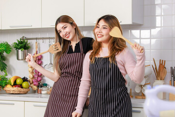 Two happy young Asian female friends, woman wearing aprons at front counter, holding a spatula feel enjoy, laughing, smiling funny on the weekend in kitchen room, Concept of teenage lifestyle at home.