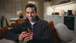© stockbusters - Smiling guy enjoy smartphone messaging sitting comfortable couch closeup.