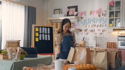 Woman preparing baked goods in cozy kitchen setup, surrounded by bread and paper bags, ready for delivery or sale.