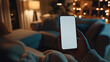 © Nayan - A close-up of a mobile phone being held by a person in a home living room. The blank white screen is clearly visible, with cozy home decor in the background, featuring soft lighting from a floor lamp