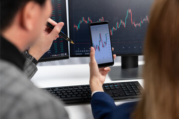 Cropped pointing hand with pen in businessman, checking with holding smartphone by woman investor online website program application with dynamic stock market at home office at back side. Infobahn.