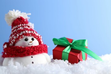 Wall Mural - Cute decorative snowman and gift box on artificial snow against light blue background, closeup