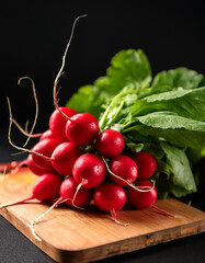 A bunch of red radishes with fresh, fresh green leaves placed on a cutting board on a black background