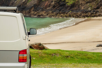 Wall Mural - Rear of a small camper van, stunning sandy beach and rock shore coastline in the background. Travel and tourism industry. Vacation on wheels theme. Journey to nature on holiday. Rented motorhome