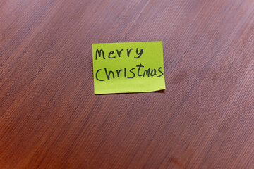 sticky note on a wooden table desk background. written note text is merry christmas