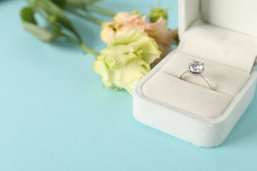 Wall Mural - Beautiful ring with gemstone in box and flowers on light blue background, closeup. Space for text