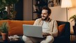 © Iona - A bearded man sitting on a cozy couch, smiling during a video call on his laptop in a warmly-lit living room.