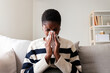 © Daniel - Sick Black woman with the flu blowing nose with tissue paper at home sitting on the sofa.