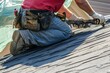 © gdgaffar - A roofer working on the roof of an apartment building, wearing work and gloves while using tools to lay shingle or tarp with a beige color background. close-up view. Generative AI