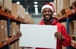 © Анастасия Терентьева - A cheerful warehouse worker in a festive Santa hat holds a blank white sign in front of a background filled with packages ready for delivery. The man is smiling warmly, ready for the holiday rush