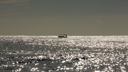 Silhouetted solo whale watching tour boat on horizon waiting for snorkelers to return from ocean with glistening waves in Moorea, French Polynesia