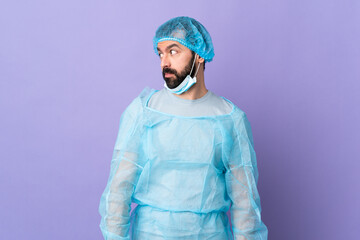 Wall Mural - Surgeon man with beard with blue uniform over isolated purple background making doubts gesture looking side