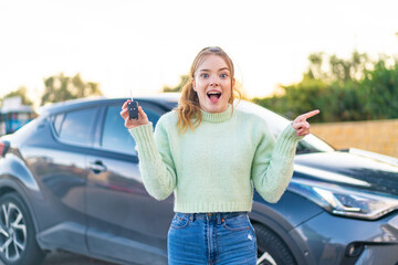 Poster - Young pretty girl holding car keys at outdoors surprised and pointing finger to the side