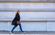 © Jacek Chabraszewski - Young blonde woman in black coat, jeans, cream turtleneck, leather handbag walking along sidewalk in city against background of gray building in autumn. Everyday life in city. Side view.