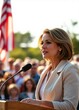 © abu - A beautiful female politician in formal attire speaking on a podium in front of a crowd, with a large American flag waving in the background.