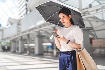 Wall Mural - Business asian woman holding UV protection umbrella with shopping bags using smartphone on sunny day